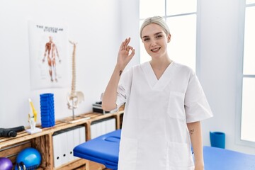 Fototapeta premium Young caucasian woman working at pain recovery clinic smiling positive doing ok sign with hand and fingers. successful expression.