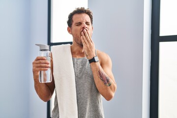 Young hispanic man wearing sportswear drinking water bored yawning tired covering mouth with hand. restless and sleepiness.