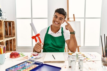 Young hispanic man at art studio holding degree smiling pointing to head with one finger, great idea or thought, good memory
