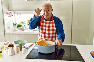 Senior man with grey hair cooking spaghetti at home kitchen annoyed and frustrated shouting with anger, yelling crazy with anger and hand raised
