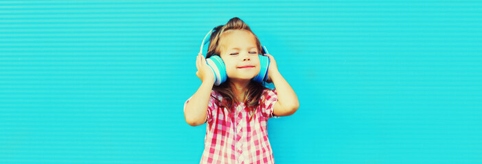 Portrait of happy little girl child in wireless headphones listening to music on blue background,...