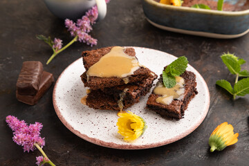 Chocolate brownie with caramel sauce with a cup of coffee on black concrete, side view.