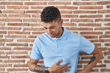Brazilian young man standing over brick wall with hand on stomach because indigestion, painful...