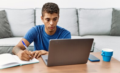 Young hispanic man studying with notebook and laptop at home