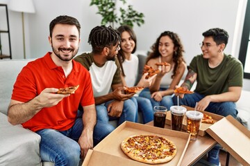 Group of young friends smiling happy eating italian pizza sitting on the sofa at home.