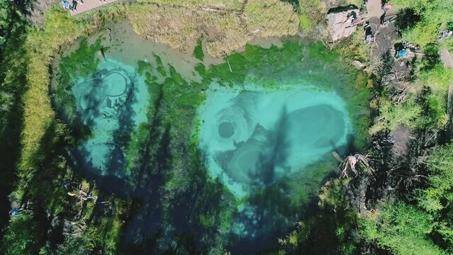 Aerial over the turquoise geyser lake with transparent mineral water and blue clay on the lakebed. Lake surrounded with green forest. Beauty in nature