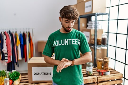 Young arab man wearing volunteer t shirt at donations stand checking the time on wrist watch, relaxed and confident