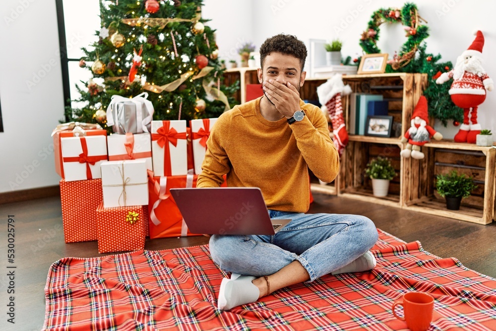 Poster Arab young man using laptop sitting by christmas tree laughing and embarrassed giggle covering mouth with hands, gossip and scandal concept