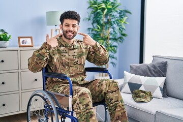 Fototapeta premium Arab man wearing camouflage army uniform sitting on wheelchair smiling cheerful showing and pointing with fingers teeth and mouth. dental health concept.