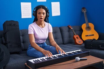 African american woman with braids playing piano keyboard at music studio scared and amazed with...