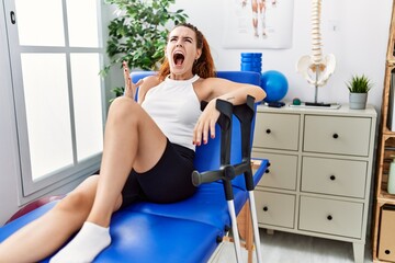 Young redhead woman lying on rehabilitation bed holding crutches crazy and mad shouting and yelling with aggressive expression and arms raised. frustration concept.