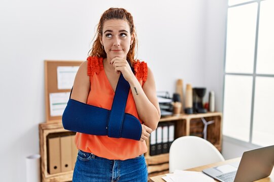 Young redhead woman wearing arm on sling at the office serious face thinking about question with hand on chin, thoughtful about confusing idea