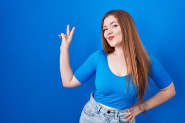 Fototapeta premium Redhead woman standing over blue background smiling looking to the camera showing fingers doing victory sign. number two.