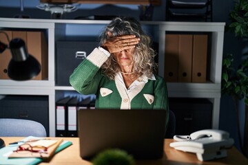 Middle age woman working at night using computer laptop covering eyes with hand, looking serious...