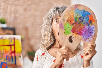 Middle age woman holding painter palette covering face afraid and shocked with surprise and amazed expression, fear and excited face.