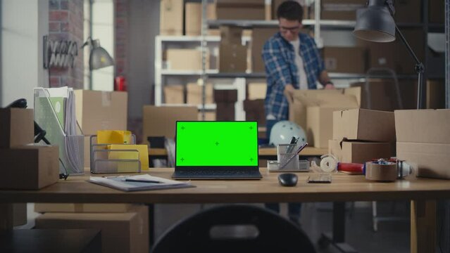 Laptop Computer Standing on a Table with a Green Screen Chromakey Mock Up Display. Small Business Warehouse with Worker Walking in the Background. Desk with Cardboard Boxes. Zoom In Shot.
