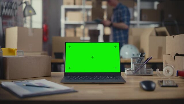 Laptop Computer Standing on a Table with a Green Screen Chromakey Mock Up Display. Small Business Warehouse with Worker Walking in the Background. Desk with Cardboard Boxes. Static Close Up Shot.