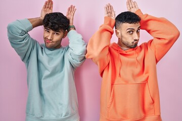 Young hispanic gay couple standing over pink background doing bunny ears gesture with hands palms...