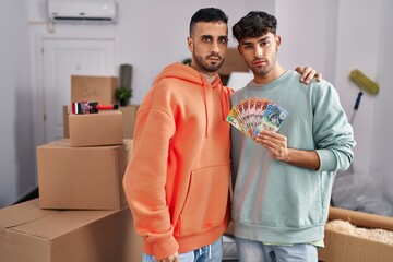 Young hispanic gay couple moving to a new home holding banknotes thinking attitude and sober expression looking self confident