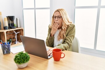 Beautiful blonde woman working at the office with laptop bored yawning tired covering mouth with...