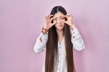 Chinese young woman standing over pink background trying to open eyes with fingers, sleepy and tired for morning fatigue