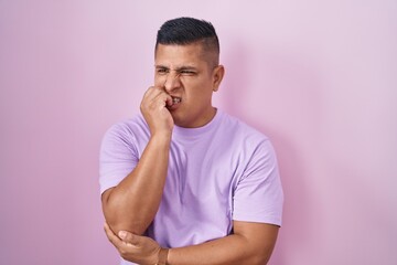 Young hispanic man standing over pink background looking stressed and nervous with hands on mouth biting nails. anxiety problem.