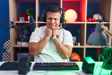 Young hispanic man playing video games laughing nervous and excited with hands on chin looking to the side