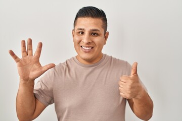 Fototapeta premium Hispanic young man standing over white background showing and pointing up with fingers number six while smiling confident and happy.