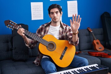 Young hispanic man playing classic guitar at music studio with open hand doing stop sign with serious and confident expression, defense gesture