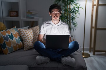 Young hispanic man using laptop at home at night puffing cheeks with funny face. mouth inflated with air, crazy expression. - obrazy, fototapety, plakaty