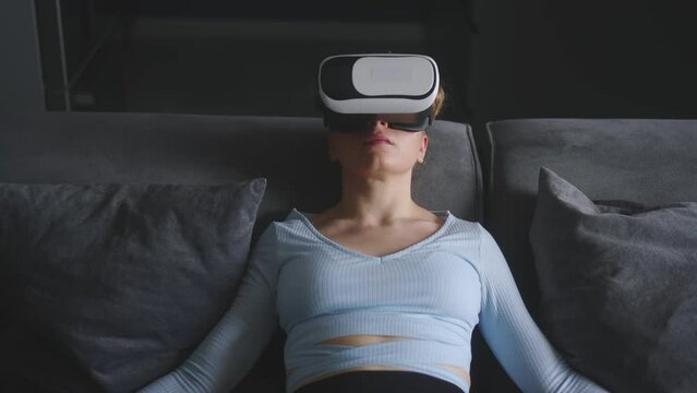 Woman in virtual reality goggles enters metaverse immersive experience via headset interface resting on sofa in minimalistic interior. Girl gaming in cyber space. Futuristic concept.