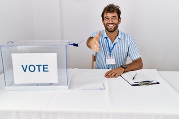 Young handsome man at political election sitting by ballot smiling friendly offering handshake as...