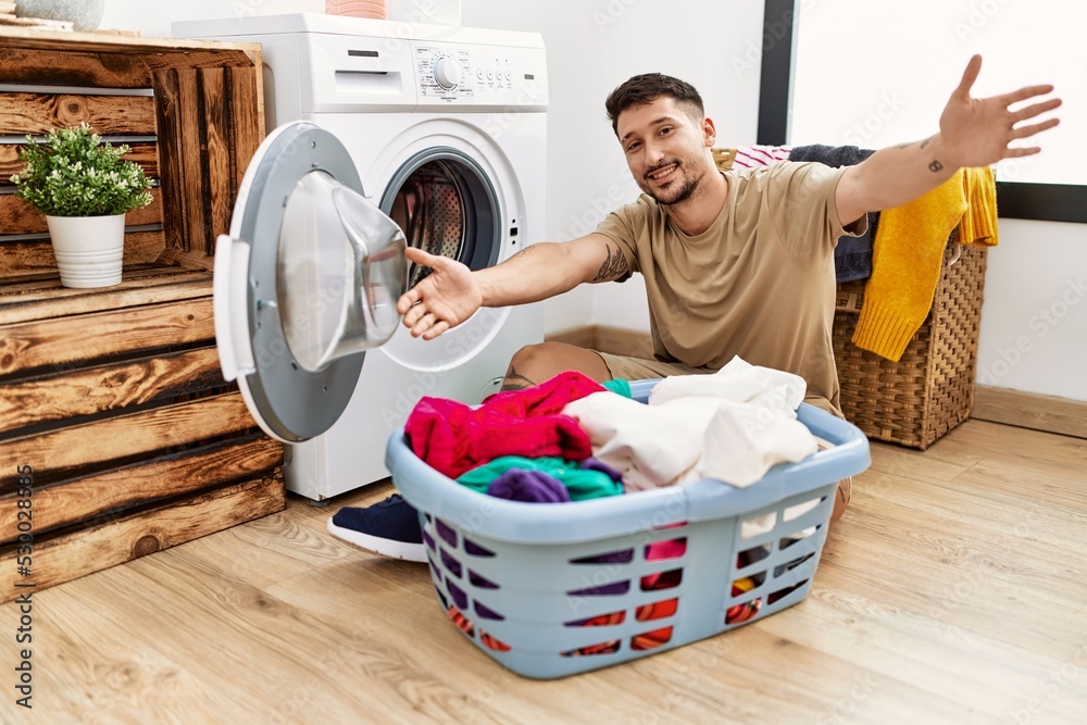 Wall mural Young handsome man putting dirty laundry into washing machine looking at the camera smiling with open arms for hug. cheerful expression embracing happiness.