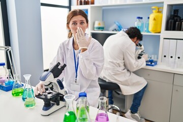 Young two people working at scientist laboratory covering mouth with hand, shocked and afraid for...