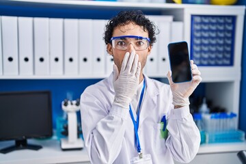 Hispanic man working at scientist laboratory showing smartphone screen covering mouth with hand,...