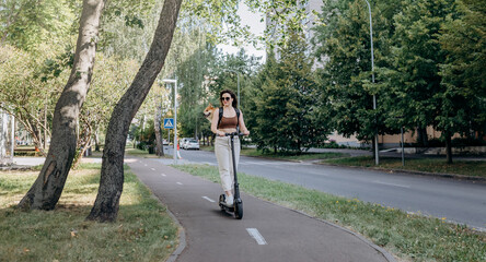 Happy smiling woman traveler is riding her electro scooter in city parkland with dog Welsh Corgi Pembroke in a special backpack