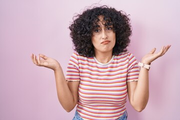 Young middle east woman standing over pink background clueless and confused expression with arms...