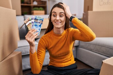 Fototapeta na wymiar Young caucasian woman sitting on the floor at new home holding canadian money stressed and frustrated with hand on head, surprised and angry face