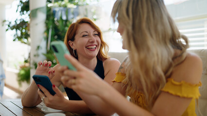 Two women using smartphones and drinking coffee sitting on table at home terrace