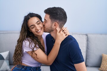 Naklejka premium Young hispanic couple sitting on sofa hugging each other and kissing at home