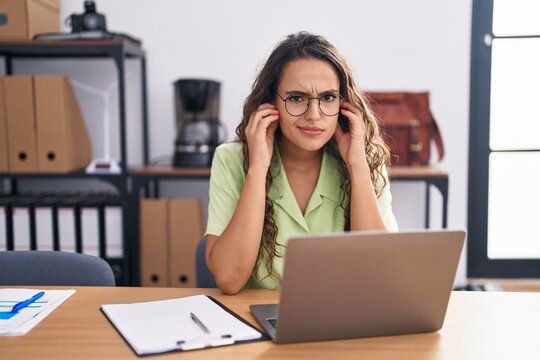 Young hispanic woman working at the office wearing glasses covering ears with fingers with annoyed expression for the noise of loud music. deaf concept.