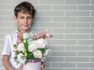 Portrait of a smiling boy in a white shirt against a gray brick wall. White Eastern European boy 9 years old with a bouquet of flowers in his hands with copy space