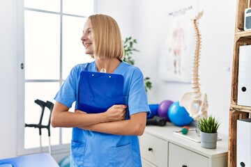 Young blonde woman wearing physiotherapist uniform holding clipboard at rehab clinic