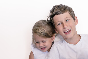A laughing brother and a shy sister stand in white clothes against a white wall. Portrait of a white Eastern European boy 9 years old and a girl 2 years old with copy space