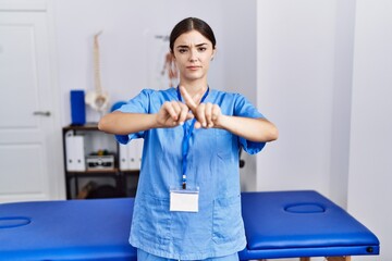 Young hispanic woman wearing physiotherapist uniform standing at clinic rejection expression crossing fingers doing negative sign - obrazy, fototapety, plakaty