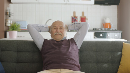 Elderly man slumping into armchair and clasping his hands behind his head in his peaceful home....