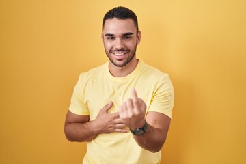 Young hispanic man standing over yellow background beckoning come here gesture with hand inviting welcoming happy and smiling