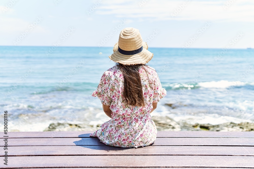 Sticker young latin girl on back view wearing summer hat sitting on the bench at the beach.