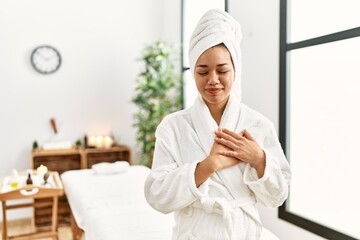 Young brunette woman wearing towel and bathrobe standing at beauty center smiling with hands on chest with closed eyes and grateful gesture on face. health concept.