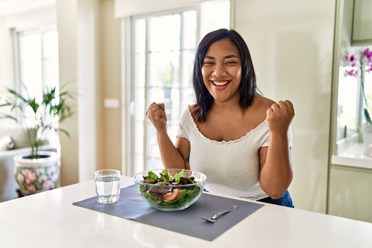 Young hispanic woman eating healthy salad at home celebrating surprised and amazed for success with arms raised and open eyes. winner concept.